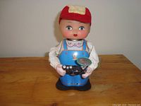 Front view of antique wind-up boy camera figurine with red cap, blue overalls, and detailed face, holding black vintage camera with flash.