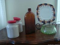 Antique whiskey bottle standing on wooden table with two white crock jugs with red lids, green depression glass bowl, and decorative square plate on stand in background.