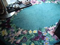 Photo of an oval hooked rug showing a teal center with detailed multicolor floral border including purple, pink, yellow, white flowers and green leaves.