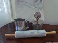 Marble rolling pin, hand mixer with glass container, and metal flour sifter arranged on table.