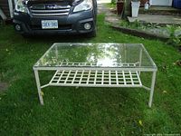 Full view of the rectangular metal patio coffee table with clear glass top and lattice shelf underneath, positioned on grass next to a vehicle.