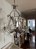 Overall view of the chandelier hanging, showing metal frame, candle-style sockets, glass/crystal drops, and chains of beads. Shows condition and missing crystals.