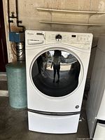 White Whirlpool front loading washing machine with broken soap dispenser drawer front, pedestal underneath, in a garage setting.