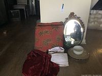 Photo showing the large accent pillow, oval ornate metal frame mirror, white dishware including bowl and plate, and some smaller glass items on a wooden floor.