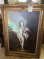 Framed oil painting of a boy holding a feathered hat, showing full front view including ornate gold frame and background scenery.