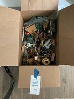 Open cardboard box filled with assorted Christmas ornaments and decorations including miniature trees, figurines, bells, and wooden items in various colors and conditions.