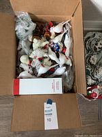 Box containing assorted Christmas tree ornaments including white birds, snowman figure, and a box with red and gold ball ornaments.