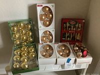 Photo showing 8 boxes of Christmas tree ornaments on a white surface next to wall, including gold and bronze baubles, Santa Claus figurines, and reindeer decoration.