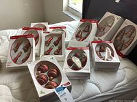 11 boxed sets of Christmas tree ornaments on bed and chair, showing variety including balls, bells, and drop shapes in red, gold, and brown colors.
