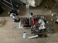 Top-down view of a table with multiple cooking utensils separated in white trays and scattered on the floor around the trays. Includes peelers, ladles, scissors, whisks, measuring spoons, and more.