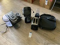 Five kitchen appliances arranged in a row on wooden floor, showing the coffee makers, blender, toaster and griddle.