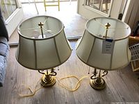 Pair of two metal base lamps with cream fabric shades placed on a wooden floor. One lamp has a yellow power cord attached, the other has none visible.