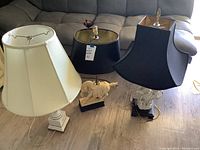 Three lamps placed on a wooden floor in front of a sofa including one white lamp with a white hexagonal shade, one black lamp with a black square shade and horse figurine base, and one black lamp with an Asian deity figurine base.