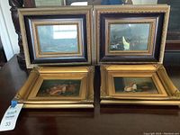 Four framed pictures displayed on table, two landscape scenes in larger black and gold frames, two small paintings of rabbits in ornate gold frames.