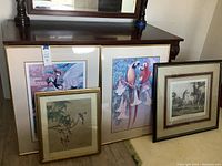 Five framed artworks leaning against a wooden dresser, two large bird prints and three smaller framed prints including one horse scene.