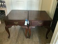 Pair of wooden end tables side by side on floor with wood-look laminate flooring, showing top view and front drawer with metal handle.