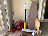 Photo showing three vacuum cleaners (red, red canister, yellow), gray ironing board, wooden chair, and blue and white iron on chair