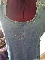 Close-up of the sleeveless denim overdress showing the neckline, button straps, and front embroidery design.