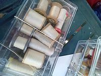 Clear plastic container with various vintage thread spools in white and beige colors, some wooden spools visible, brand markings partially visible.