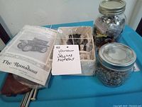 Photo showing needlecraft pattern and jars with buttons and pins alongside a box of assorted sewing notions.