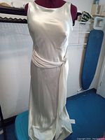 Front view of vintage white satin formal gown on dress form showing silky texture, sleeveless design, and oblique draped buckle detail on waist.