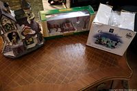 Photo showing the Santa's Workshop building illuminated with adapter, the Maple Syrup Run set in box, and the Christmas Street ice sculptors boxed accessory.