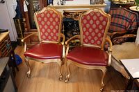 Two matching gold French style chairs with casters, red patterned fabric backs and red leather seats, displayed on wooden floor.