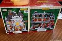 Two boxed LEMAX Village Collection lighted buildings including Village Fire House No. 1 and Canadian Tire building showing front viewing of packaging.