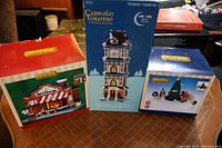Three boxed items on table: LEMAX Posh Pets Salon, Carole Towne Big Carole Clock Tower, and Skateboard Mushers accessory boxes, showing packaging and brands.