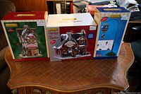 Photo of all three boxed LEMAX village buildings and one animated accessory together on a wooden table.
