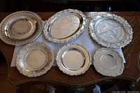 Six small silver plated serving trays arranged on a table showcasing various shapes and ornate borders.