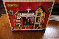 Photo of the front of the LEMAX Rising Star Christmas Cookies factory box showing detailed artwork of the product, featuring a festive miniature bakery building with Christmas themed decorations and signage.