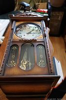 Wooden wall clock showing full frontal view with detailed lower door and two pendulums visible.