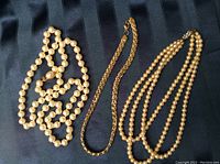 Photo showing three necklaces on a dark fabric background: one single strand knotted pearl necklace with gold clasp, one triple strand pearl necklace, and one woven gold chain necklace.