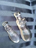 Photo showing two clear glass hippo figurines side by side on a dark striped fabric background, one larger and one smaller.