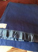 Photo showing the folded marine blue Merino wool blanket with satin trim edge and partial label visible.