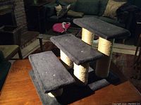 Three-step cat climber with black carpeted steps and beige sisal-wrapped cylindrical posts shown on a wooden surface with couch and fireplace background.