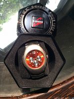 Metal hexagonal G-Shock branded case with the watch inside showing red dial and black strap.
