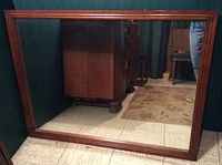 Front view of the large rectangular mirror showing the reddish hardwood frame and clear reflective surface, placed upright on floor against a dark-colored wall.