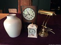 Overall group photo showing the wooden framed Hamilton 31 day mantle clock, large white porcelain vase, porcelain floral motif desk clock, and brass desk lamp on a dark purple textured surface.