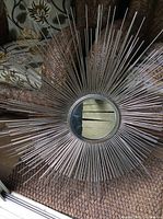 Front view of the large circular sunburst mirror with metallic radial rods around the central glass mirror, sitting on a carpeted floor.