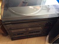 Photo of Toshiba sound system showing vinyl record player with dust cover on top, stacked CD and DVD player units below, placed on wooden floor.