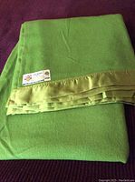 Folded emerald green 100% Merino wool blanket with a green satin trim and a tag visible on the corner.