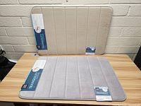 Two bathroom mats, beige and light gray, shown flat on a wooden surface with labels visible.