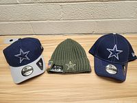 Photo showing two Dallas Cowboys caps and one green knit beanie, all with team star logo visible, on wooden surface against brick wall.