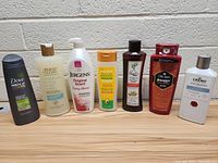 Image showing seven assorted full-sized bottles of personal care products including shampoos, body washes, and lotions lined up on a wooden surface against a white wall.