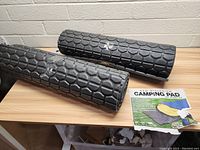 Two rolled black geometric design foam camping pads on a wooden table with the promotional sheet.