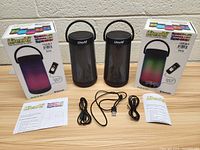 Two black LitezAll wireless LED speakers, each accompanied by a remote control, USB charging cable, auxiliary charging cable, user manuals, and their retail boxes.