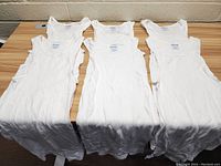 Six white men's undershirts laid flat on a wooden surface, showing front view and tags, arranged in two rows of three.