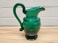 Front view of the green handblown glass pitcher on a wooden surface against brick wall background.